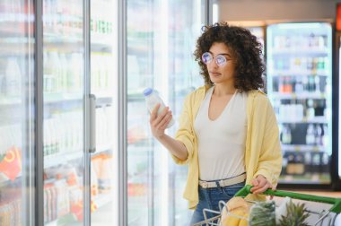 Genç bir kadın marketin soğutulmuş bölümünden bir şişe süt seçiyor, alışveriş arabasını itiyor.