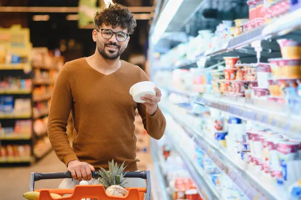 Müşteri dondurucu koridordan yoğurt seçerken açık ve neşeli bir süpermarkette içi yiyecek dolu bir alışveriş arabasını itiyor.