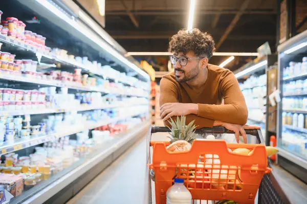 Market alışverişi yapan, alışveriş arabasına yaslanan ve dondurulmuş süt ürünlerine bakan genç bir adam.