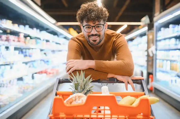 Genç Arap adam markette alışveriş yaparken alışveriş arabasında gülümsüyor ve yaslanıyor.