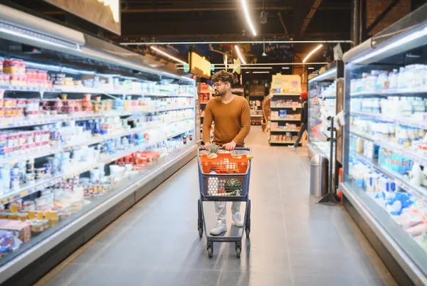 Genç Arap adam marketten alışveriş arabası alıyor, dondurucu koridorlarda yürüyor.