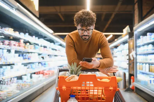 Süpermarkette alışveriş yaparken akıllı telefonuna danışan, alışveriş listesini kontrol eden ya da anlaşma arayan bir adam.