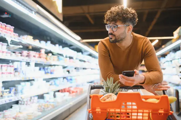 Genç adam akıllı telefon kullanıyor. Parlak, düzenli bir süpermarkette alışveriş yapıyor, fiyatları kontrol ediyor ve yeni ürünler seçiyor.