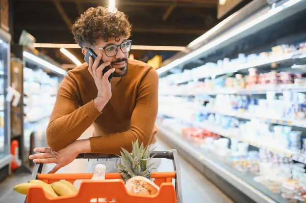 Genç bir adam markette alışveriş yaparken telefonla konuşuyor. Yeni ürünler seçiyor ve alışveriş arabasını dolduruyor.