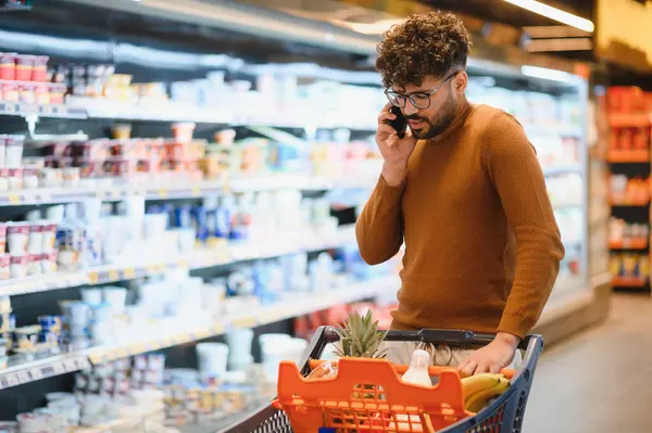 Müşteri, kalabalık bir süpermarketin içinde, koridorlarda ve raflarda gezinirken bir yandan bakkaldan yiyecek seçiyor.