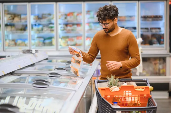 Müşteri süpermarkette geziniyor, dondurulmuş bir ürünü elinde tutarken market poşetleriyle dolu bir alışveriş arabasını itip etiketleri kontrol ediyor.