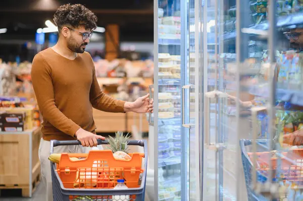 Müşteri bir süpermarkette buzdolabının kapısını açıyor, taze yiyecek ararken parlak koridorda bir alışveriş arabasını itiyor.