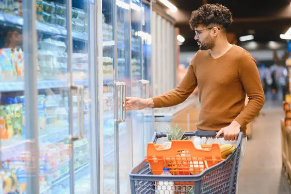 Genç Arap adam süpermarkette buzdolabı açarken market alışverişi yapıyor, alışveriş arabası itiyor.