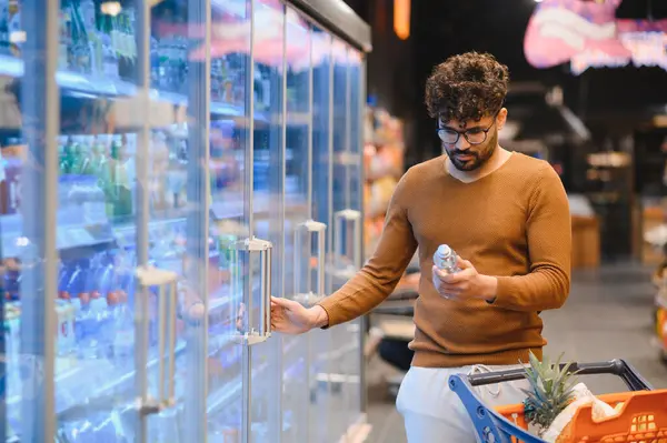 Müşteri marketin soğutuculu koridorunda yiyecek seçiyor, şişelenmiş su etiketi okuyor.