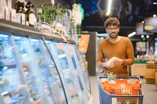 Genç adam market alışverişi yapıyor, alışveriş arabası itiyor ve süpermarketin buzlu bölümünden peynir seçiyor.