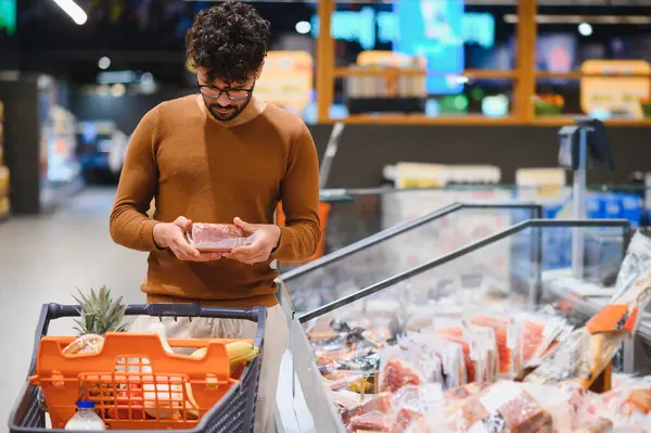 Müşteri marketteki buzdolabından paketlenmiş et seçiyor, alışveriş arabasıyla alışveriş yapıyor.