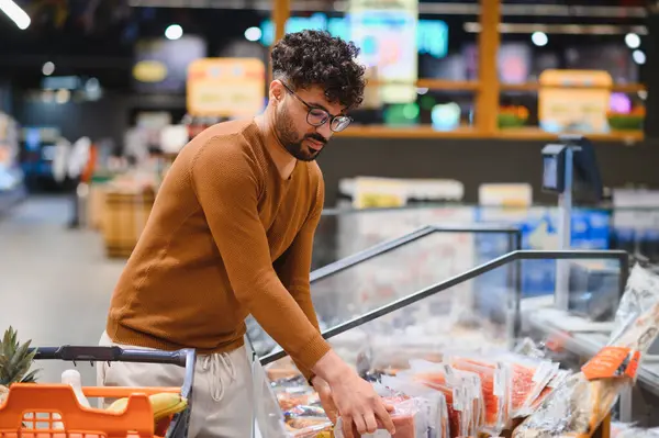 Genç Arap adam marketten alışveriş yapıyor soğutulmuş bölümden doğranmış jambon seçiyor.
