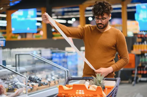 Genç adam uzun bir market fişini incelerken bir süpermarketin koridorlarında geziniyor, harcamaları ve bütçeyi etkin bir şekilde yönetiyor.