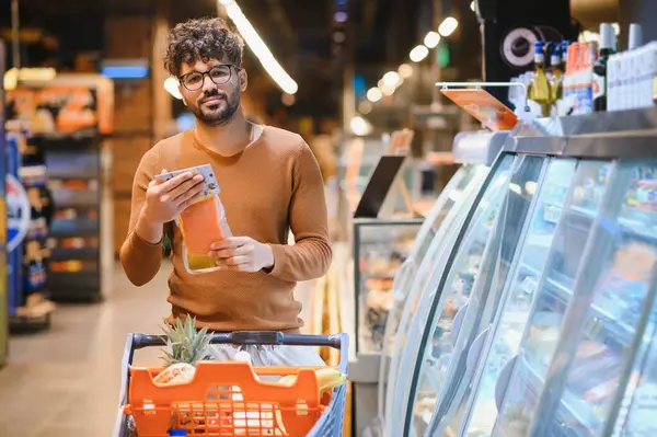 Genç adam market alışverişi yapıyor, marketin buzdolabı koridorunda somon paketi etiketi okuyor.