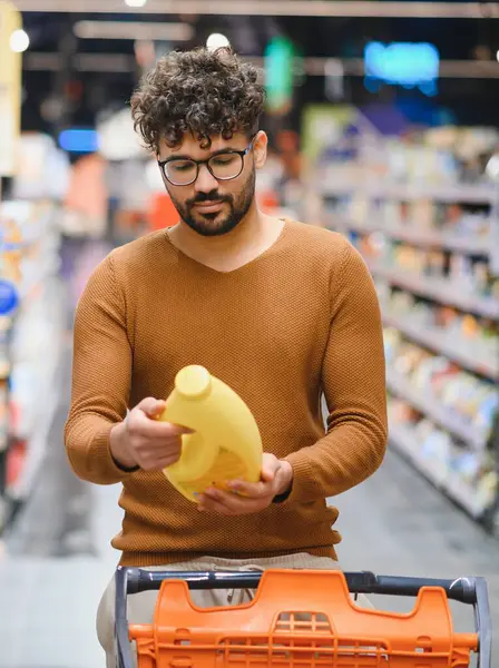 Odaklanmış Arap adam markette alışveriş yaparken temizlik malzemelerini dikkatlice kontrol ediyor.