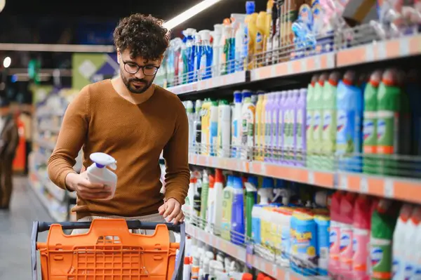 Odaklanmış Arap adam süpermarkette temizlik ürünlerini seçiyor, alışveriş arabasını itiyor ve deterjan şişesinde etiket okuyor.