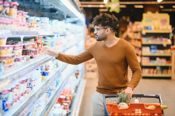 Genç Arap adam süpermarkette alışveriş yapıyor, dondurulmuş süt bölümünden yoğurt seçiyor, alışveriş arabasını itiyor.