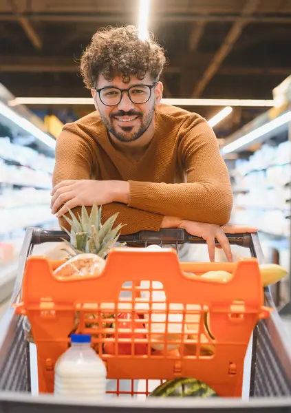 Alışveriş arabasının üzerine yaslanmış, markette taze yiyeceklerle dolu mutlu bir müşteri.