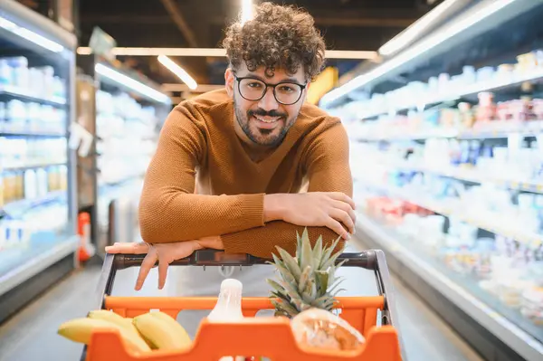 Genç Arap adam, canlı bir süpermarket koridorunda alışveriş arabasına yaslanırken gülümsüyor.
