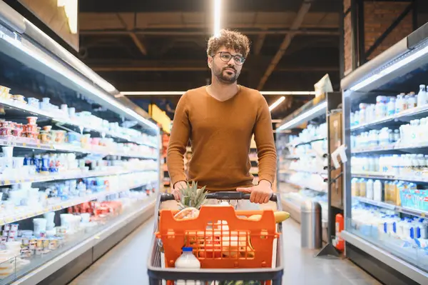Müşteri, açık, düzenli bir süpermarket koridorunda alışveriş arabasını itiyor, günlük ihtiyaçlar için taze yiyecek ve gereçleri seçiyor.