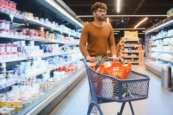 Genç bir Arap, alışveriş arabasını parlak bir süpermarket koridorunda itiyor, taze yiyecek seçiyor ve tatmin edici bir alışveriş deneyiminin tadını çıkarıyor.