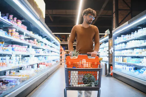 Genç bir Arap süpermarkette alışveriş arabasını itiyor, dondurucu koridorlarda yürüyor.