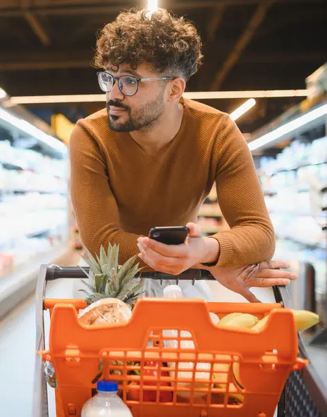 Genç bir adam markette alışveriş arabasına yaslanırken akıllı telefonuna danışıyor.
