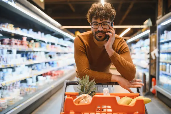 Genç adam marketten alışveriş yaparken akıllı telefon kullanıyor, marketten sağlıklı yiyecek seçiyor.