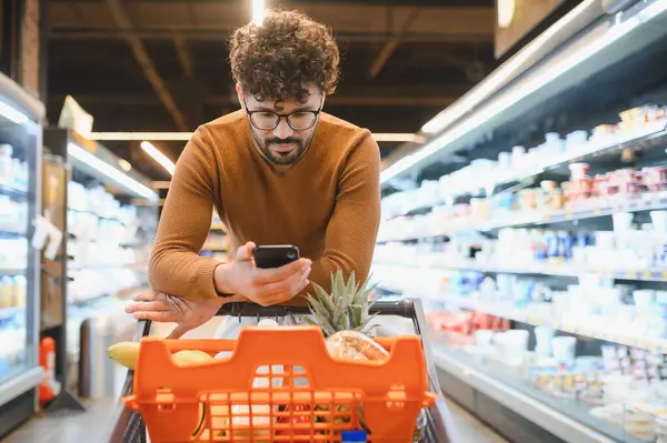 Genç adam alışveriş listesini akıllı telefondan kontrol ederken marketin kalabalık koridorunda taze yiyecek seçiyor.