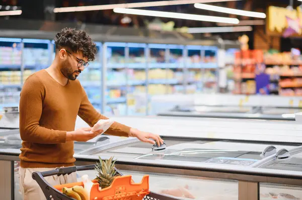 Müşteri markette alışveriş yaparken yiyecek etiketlerini analiz ediyor, soğutulmuş bölümden dondurulmuş ürünleri dikkatlice seçiyor.