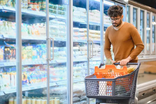 Odaklanmış Arap adam alışveriş arabasını iterken kalabalık bir süpermarket koridorunda taze yiyecekler seçiyor ve sağlıklı bir yaşam tarzının tadını çıkarıyor.