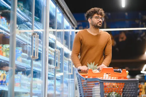 Müşteri bir alışveriş arabasını süpermarket koridorunda itiyor, canlı perakende ortamında gezinirken taze yiyecek seçiyor.