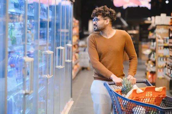 Genç Arap adam alışveriş arabasını itiyor ve süpermarketteki cam kapılı buzdolabından donmuş gıdayı seçiyor.