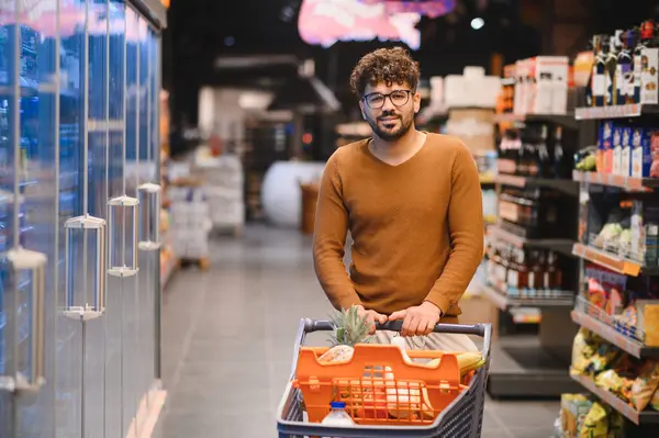 Genç bir adam markette alışveriş arabasını itiyor, soğutulmuş rafların yanında yürüyor.