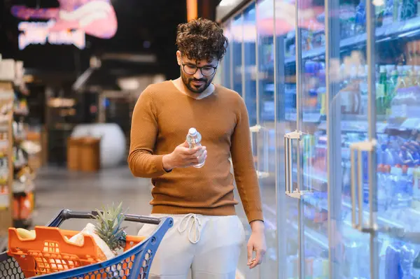 Genç bir adam alışveriş arabasını itiyor ve süpermarket buzdolabı bölümünden bir şişe su seçiyor.