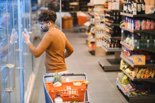 Arap müşteri süpermarkette soğutuculu koridordan bir şişe su seçiyor, market alışverişi yapıyor.