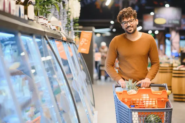 Arap bir adam alışveriş arabasını parlak bir süpermarket koridorunda itiyor, taze yiyecek seçiyor ve modern bir alışveriş deneyiminin tadını çıkarıyor.
