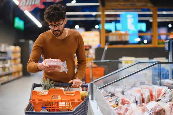 Genç Arap adam alışveriş arabasını itiyor ve marketteki paketlenmiş etin son kullanma tarihini kontrol ediyor.