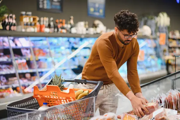Genç adam market alışverişi yapıyor. Parlak ışıklı bir süpermarkette dondurulmuş etten paketlenmiş et seçiyor.
