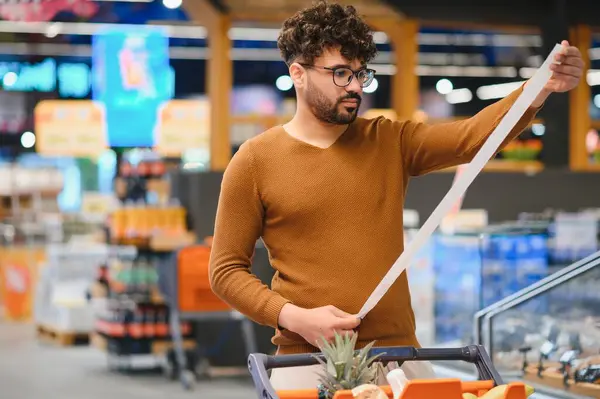 Genç adam süpermarkette alışveriş yaparken çok uzun bir market fişini dikkatle inceliyor ve artan gıda maliyetleri hakkındaki endişelerini dile getiriyor.