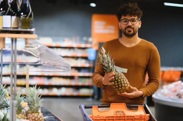 Markette ananas tutan, alışveriş arabasıyla yürüyen genç bir adam.