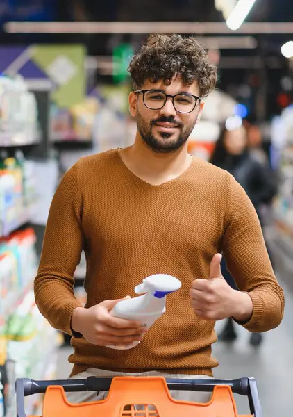 Market alışverişi yapan genç adam temizlik ürünlerini tutuyor ve süpermarkette memnuniyet belirtileri gösteriyor.