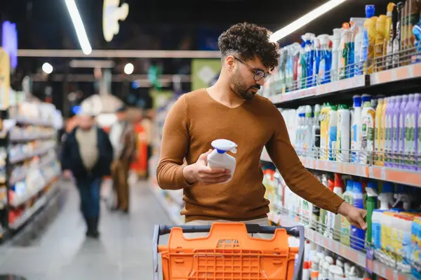Müşteri raftan temizlik malzemesi seçerken, alışveriş arabasını kalabalık bir süpermarket koridoruna itiyor.