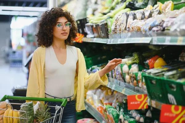 Genç bir kadın süpermarkette market raflarını karıştırıyor, yeni ürünler ve alışveriş arabası için sağlıklı seçenekler seçiyor.