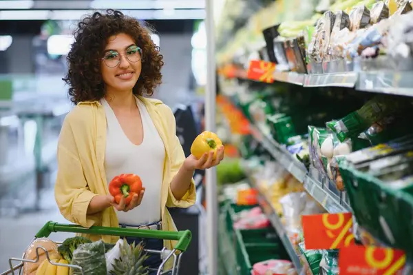 Markette alışveriş yaparken biberleri karşılaştıran, elinde alışveriş arabası olan genç bir kadın.