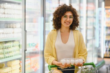 Süpermarketin buzdolabı reyonunda alışveriş yaparken elinde bir paket yumurta tutan gülümseyen kadın.