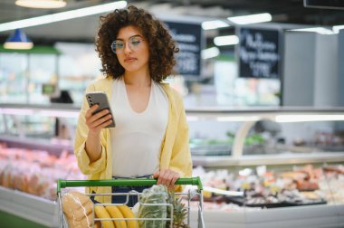 Markette alışveriş arabasını iterken akıllı telefon kullanan, alışveriş listesini kontrol eden ya da fiyatları karşılaştıran genç bir kadın.