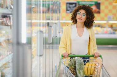 Müşteri alışveriş arabasını itiyor ve marketin buzdolabı bölümündeki dondurulmuş yiyecekleri seçiyor.