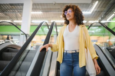 Bir markette yürüyen merdivene çıkan genç bir kadın, içi taze ve sağlıklı bakkaliyelerle dolu bir alışveriş çantası taşıyor.