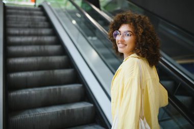 Kıvırcık saçlı ve gözlüklü genç bir kadın alışveriş merkezinde yürüyen merdivene çıkıyor ve elinde tekrar kullanılabilir bir alışveriş çantası var.